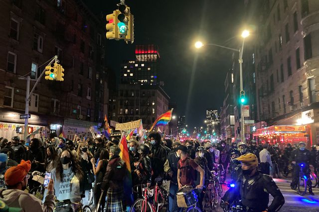 A group of trans rights protesters fill the streets waving a rainbow flag at night in downtown Manhattan.